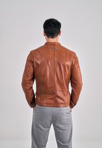 Veste en cuir marron avec un col rond et des détails de coutures, portée par-dessus un pantalon gris. Le dos comporte un petit logo embossé.