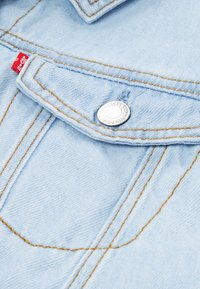 Ανοιχτό μπλε τζιν μπουφάν με αντίθετες πορτοκαλί ραφές, ασημένιο κουμπί τύπου snap και κόκκινη ετικέτα μάρκας στην τσέπη.