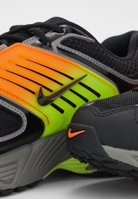 Baskets Nike noires et grises avec des panneaux latéraux orange néon et verts et petits logos Nike orange sur la semelle et la tige.