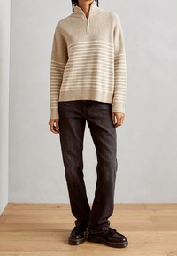 Světlý béžový svetr na zip s vertikálními pruhy, žebrovanými manžetami a lemem, spárovaný s tmavě šedými džíny s rovnými nohavicemi a černými botami.