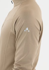 Beige langemouwen top met een hoge kraag en een gladde textuur. Beschikt over een klein blauw driehoekig logo op de linkermouw.