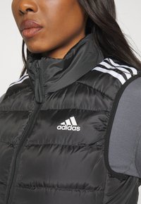 Juodas pūkinis liemenė su aukštu apykakle, užtrauktuku ir baltu adidas logotipu. Yra baltos juostelės ant pečių ir siuvinėta tekstūra.