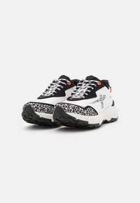 Zapatillas en blanco y negro con suela texturizada, que presentan un patrón geométrico en la puntera y cordones negros. Acabados en naranja en el talón.