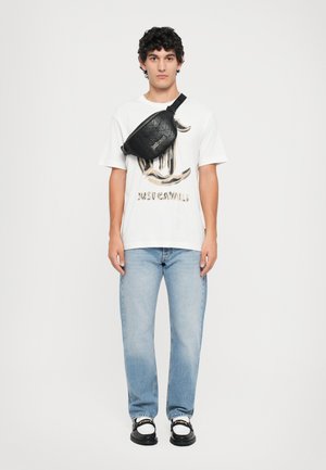 Un rucsac negru cu model embosat purtat peste un tricou alb cu grafică, ce prezintă text auriu, asortat cu blugi bleu deschis și pantofi negri.