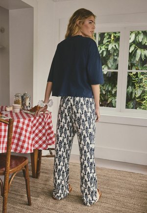 Frau steht in einer Küche, trägt eine Hose mit Fischmuster und ein marineblaues Hemd, die Hand ruht auf einer rot-weiß karierten Tischdecke.