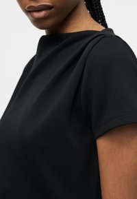 Κοντινό πλάνο ενός ατόμου που φοράει μαύρο τοπ με κοντά μανίκια και ψηλό λαιμόκοψη και ορατά πλεκτά μαλλιά.