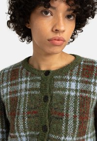 Zöld kockás kardigán, texturált kötött anyagból készült, kerek nyakú, négy fekete gombbal és vörös-fehér minták keverékével.