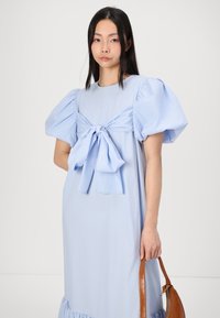 Robe bleu clair à manches bouffantes avec détail en nœud à l'avant, silhouette fluide et ourlet en volants. Modèle tenant un sac à main en cuir structuré marron.