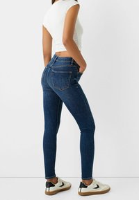 Jeans ajustados en denim oscuro con una cintura alta, con costuras sutiles, dos bolsillos traseros y un corte hasta los tobillos, combinados con zapatillas blancas.
