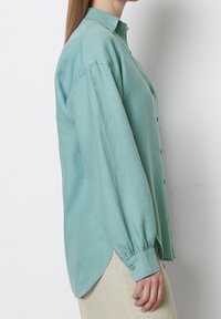 Camicia di lino blu chiaro con colletto, chiusura a bottoni sul davanti, maniche lunghe con polsini abbottonati e orlo leggermente curvato.