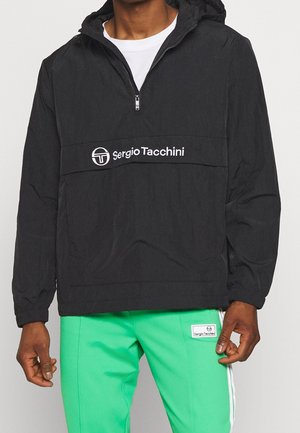Personne portant une veste à capuche noire Sergio Tacchini avec une fermeture éclair courte et un pantalon vert Sergio Tacchini avec des bandes blanches sur les côtés.