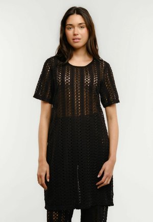 Nő fekete, rövid ujjú horgolt ruhában, fekete nadrág felett, egyszínű fehér háttér előtt állva.