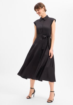 Femme portant une robe midi noire sans manches boutonnée avec une ceinture à la taille, associée à des sandales noires à talons hauts à bride cheville, souriante et regardant vers le bas.