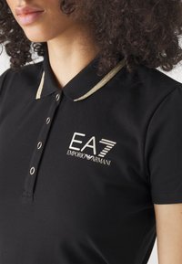 Svart pikétröja med en guldfärgad krage, tre knappslutningar och broderad logotyp med texten "EA7 Emporio Armani."