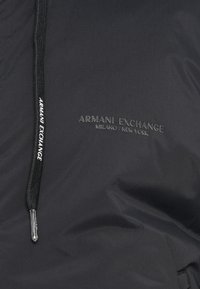Juodas audinys su išsiuvinėtu "ARMANI EXCHANGE MILANO / NEW YORK" logotipu ir juodu raišteliu, kuris puošia baltas "ARMANI EXCHANGE" tekstas.