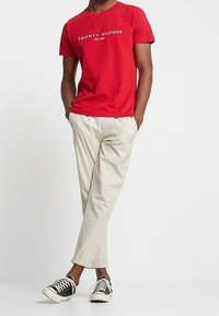 Camiseta de algodón roja con el logo "TOMMY HILFIGER", combinada con pantalones beige claros y zapatillas en blanco y negro. Conjunto casual exhibido.