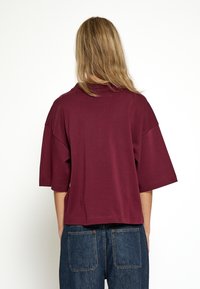 Camiseta oversized burdeos de mangas cortas, con un escote redondo y un dobladillo recto. Combinada con pantalones de mezclilla azules.