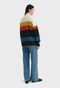 Gebreide trui met hoge kraag, voorzien van horizontale strepen in crème, geel, rood, bruin en blauw. Combinatie met gestructureerde blauwe corduroybroek.