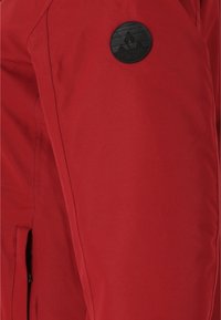 Röd vattentät jacka med en slät yta, vertikala sömmar och en rund svart logotypmed ett bergmotivet.