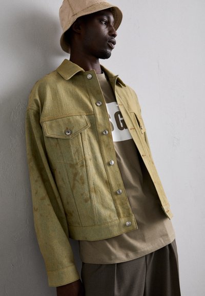 Light green denim jacket with front pockets, silver button closures, and contrasting stitching, paired with a beige t-shirt and trousers.