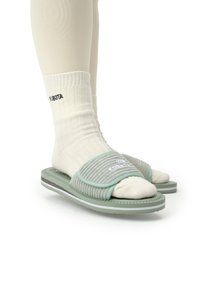 Des sandales ouvertes vert clair avec des brides grises côtelées, portées sur des chaussettes en tricot blanches. Elles possèdent une semelle texturée avec des accents blancs et gris.