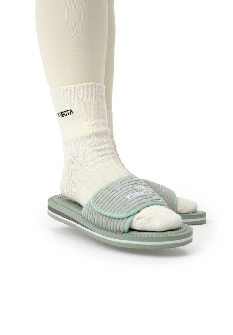 Des sandales ouvertes vert clair avec des brides grises côtelées, portées sur des chaussettes en tricot blanches. Elles possèdent une semelle texturée avec des accents blancs et gris.