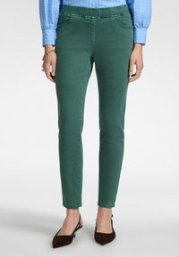 Pantalones ajustados verdes con bolsillos frontales, confeccionados en una suave mezcla de algodón. Cintura elástica, combinados con zapatos planos marrones de punta.