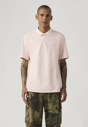 Junger Mann mit kurzen Haaren und Arm-Tattoos, der ein hellrosa Polohemd und grüne Tarn-Cargohosen trägt und vor einem schlichten Hintergrund steht.