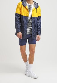 Voděodolná bunda s designem v blokových barvách žluté a námořnické, spárovaná se pruhovanou košili a námořnickými šortkami, doplněná o bílé tenisky.
