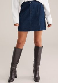 Sötét farmer mini szoknya elöl zsebekkel és gombos zárással; térdig érő, barna bőr magassarkú csizmával párosítva.