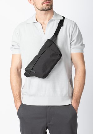 Borsa a tracolla nera in materiale liscio con forma rettangolare. Dotata di tracolla regolabile e dettaglio discreto del logo. Indossata a tracolla diagonalmente sul petto.