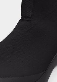 Κοντινό πλάνο μαύρου μποτάκι αστραγάλου με υφαντή υφασμάτινη επάνω επιφάνεια και λεπτομέρεια με λωρίδα τραβήγματος κοντά στον αστράγαλο, με λεία καουτσούκ σόλα.