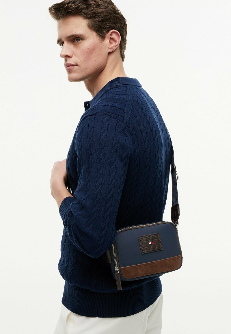 Jeune homme portant un pull torsadé bleu marine et un pantalon blanc porte un sac bandoulière bleu marine et marron avec un écusson logo.