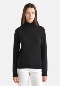 Pull à col roulé noir en tissu doux, avec des manches longues et une coupe ajustée. Porté avec un pantalon blanc, avec un minimum de détails.
