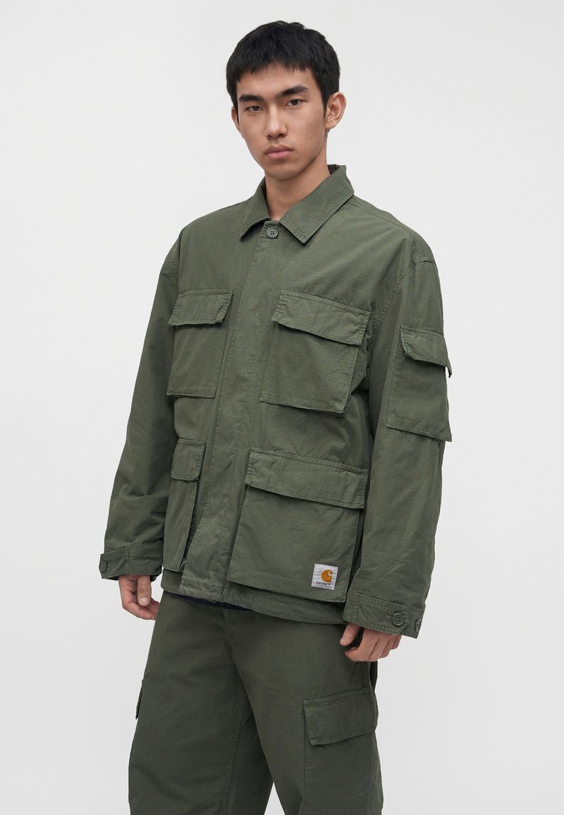 Joven con chaqueta de trabajo verde y pantalones cargo a juego, de pie frente a un fondo claro liso con expresión neutral.
