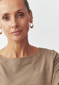 Top marron, hors des épaules avec une texture douce et des détails de couture subtils. De grandes boucles d'oreilles en or ajoutent une touche à l'encolure.