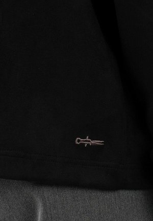 Svart stoff med en liten, lys grå brodert logo som ligner et stilisert sverd eller dolk nær kanten.