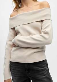 Bež pulover z odkrito rameni, rebrastim ovratnikom, slim fit silhueto, dolgimi rokavi in mehkim materialom. V kombinaciji z temnim jeansom za kontrast.