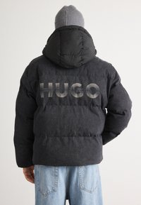 Svart pufferjakke med hette, laget av teksturert stoff, med "HUGO" logoen trykket i store, lysegrå bokstaver på baksiden.