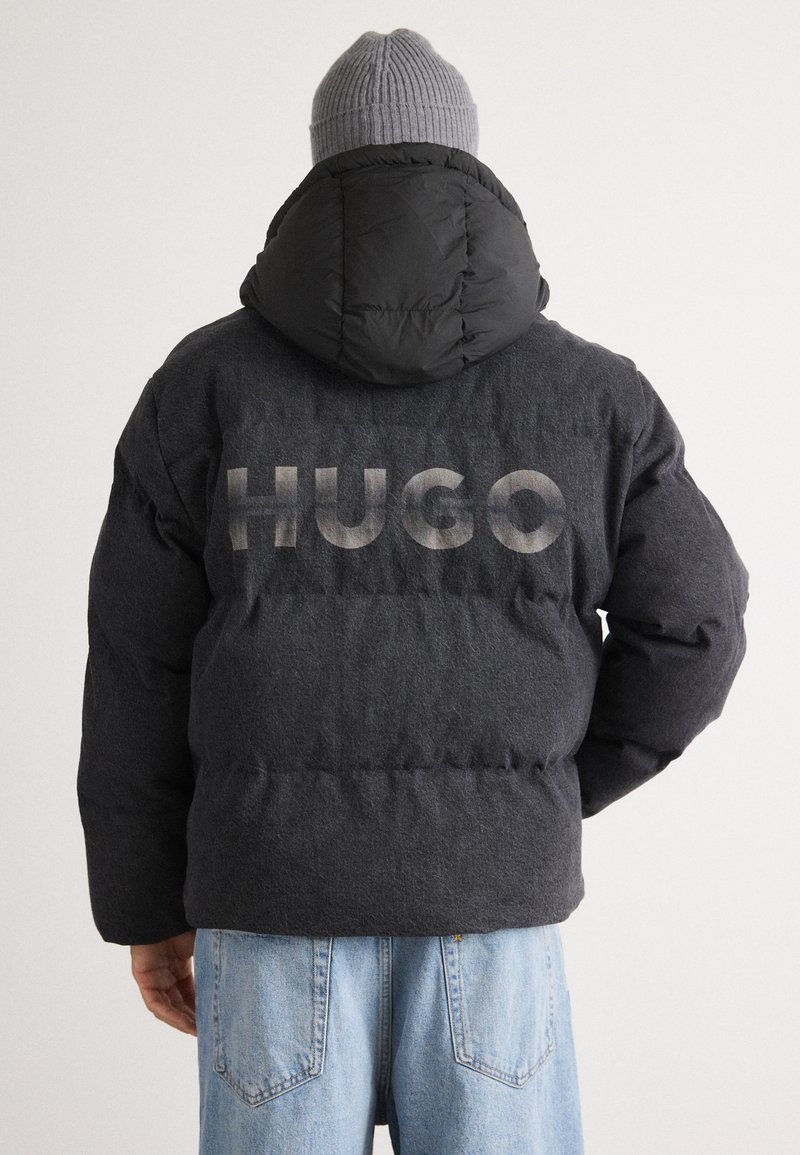 Svart pufferjakke med hette, laget av teksturert stoff, med "HUGO" logoen trykket i store, lysegrå bokstaver på baksiden.