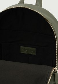 Lo zaino verde in pelle presenta un interno con una fodera in tessuto nera, un compartimento con zip e un'etichetta del marchio discreta nella tasca interna.