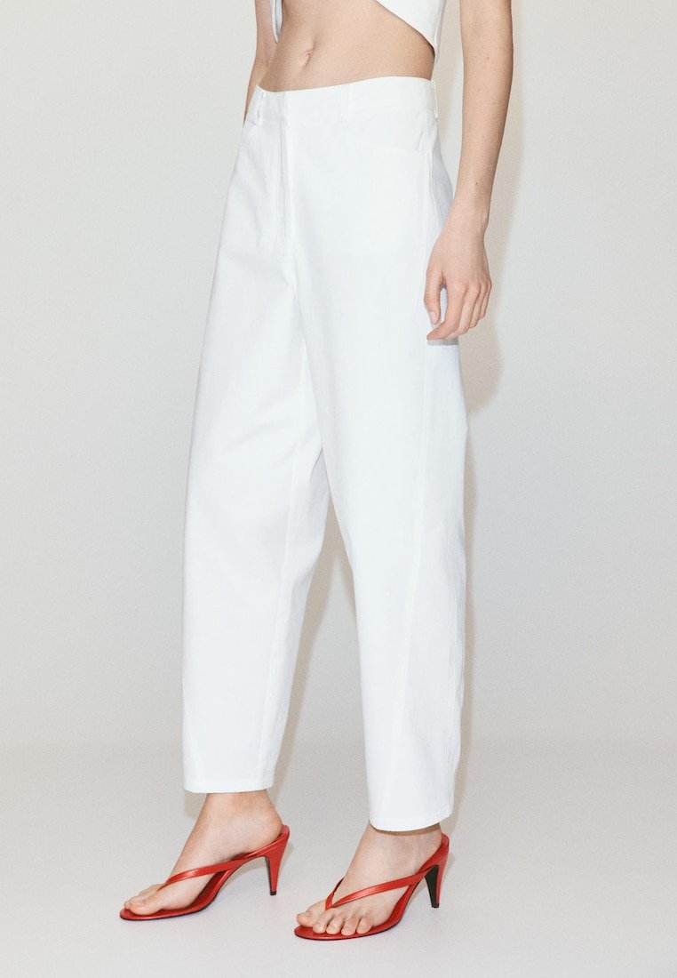 Femme portant un pantalon blanc taille haute et des sandales rouges à talons, debout devant un fond clair uni.