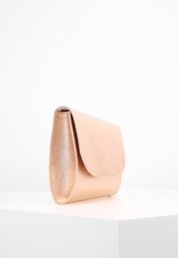 Malá metalická kabelka v odstínu růžového zlata s klopou, vystavená na bílém podstavci na jednobarevném bílém pozadí.