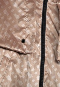 Lehké béžové sako s lesklým povrchem, které má texturovaný vzor, černý zip a detail v podobě kovového očka.