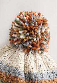 Kötött krémszínű sapka színes bojttal, amely narancs, zöld, kék és fehér árnyalatait tartalmazza, texturált, bordázott mintával.