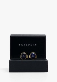 Gemelos de tono dorado con incrustaciones ovaladas de esmalte azul que presentan un diseño de calavera y huesos cruzados en blanco, presentados en una caja negra texturizada.