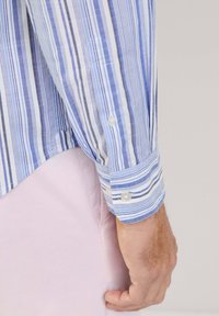 Camicia a righe con linee blu, bianche e grigie, dotata di polsini con bottone e tessuto morbido. Mano visibile che tiene il polsino.