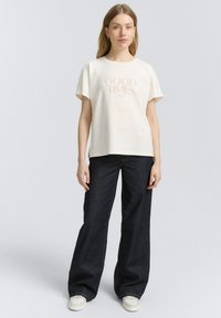 T-shirt crème à manches courtes avec le texte "GOOD TIMES" en rose clair, associé à un jean large bleu foncé et des baskets blanches. Design simple et décontracté.