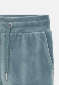 Měkké, světle modré tepláky s páskem na stažení v pase, stříbrnými kovovými kroužky a bočními kapsami; materiál vypadá plyšově a texturovaně.