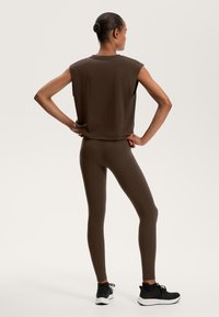 Barna ujjatlan felső lekerekített nyakkivágással, amely jól illik a szűk barnás leggingshez és fekete sportcipőkhöz. Egyszerű, sima anyag és dizájn.